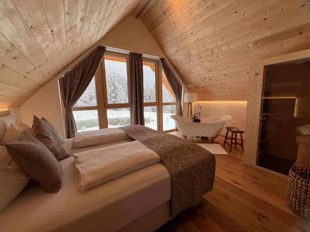 Großes Schlafzimmer in Holzhaus mit Panoramafenster, Badewanne und Winterblick in den Bergen.